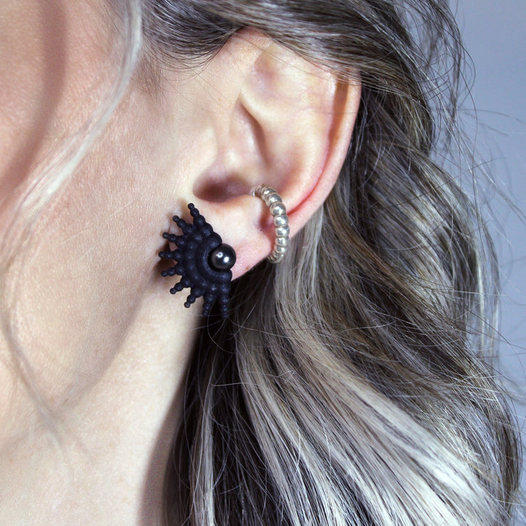 This sculptural ear cuff from the PHAOS collection reimagines the ancient granulation technique through digital design and contemporary craftsmanship. Designed to sit securely without a piercing, its fluid form curves around the ear and finishes with a luminous white glass pearl - adding a touch of shimmer to any look. Available in your choice of gold-plated brass or .925 silver.